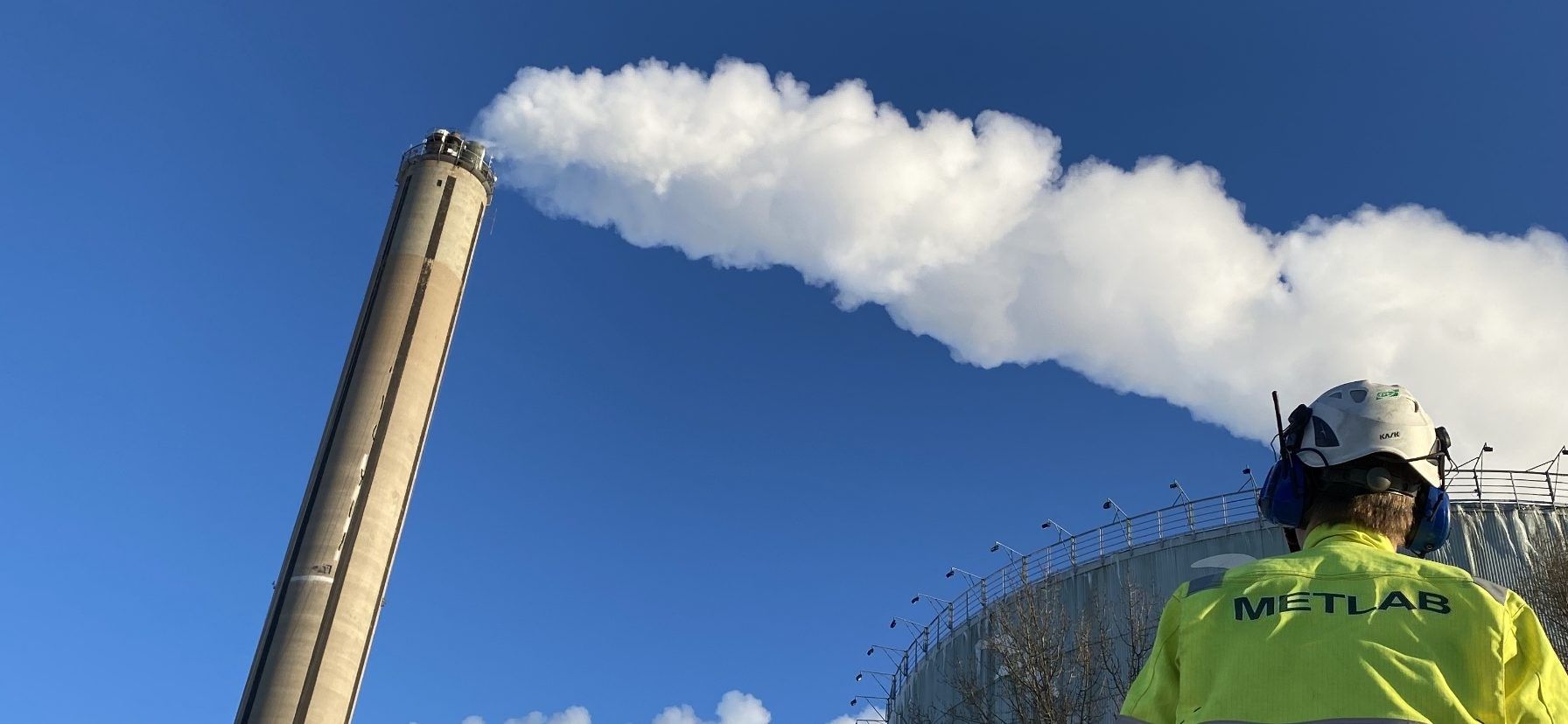METLAB-ingenjör framför en skorsten med rökgasplym mot blå himmel